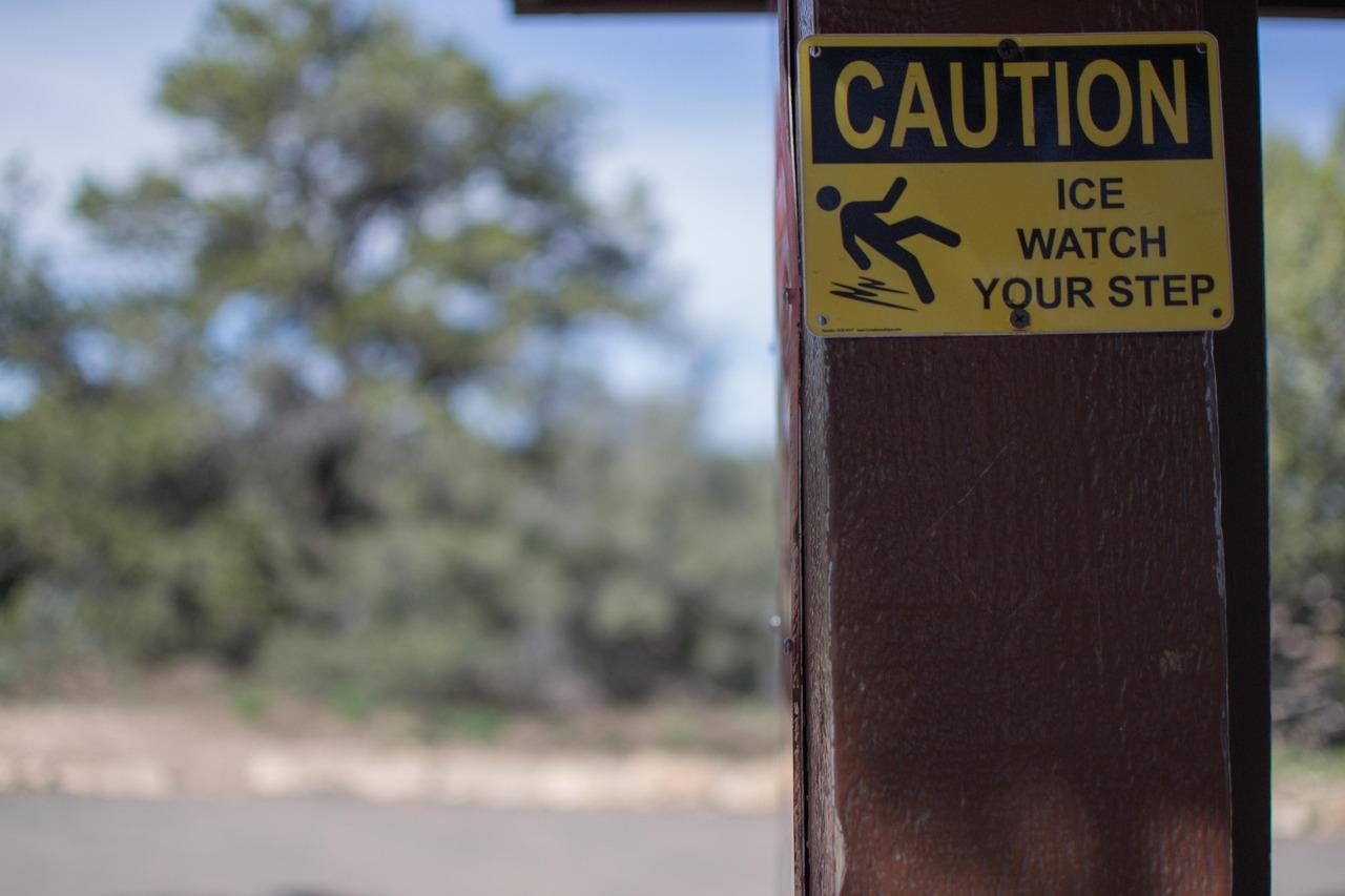 A yellow sign on a wooden post warns, “CAUTION: ICE WATCH YOUR STEP” with an icon of a person slipping. Blurred trees and a path are visible in the background.