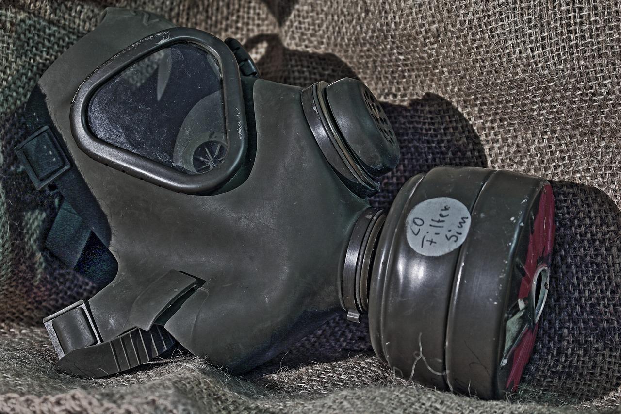 A black gas mask with a filter labeled CO filter Sim rests on rough, brown burlap fabric, reflecting light on its visor.