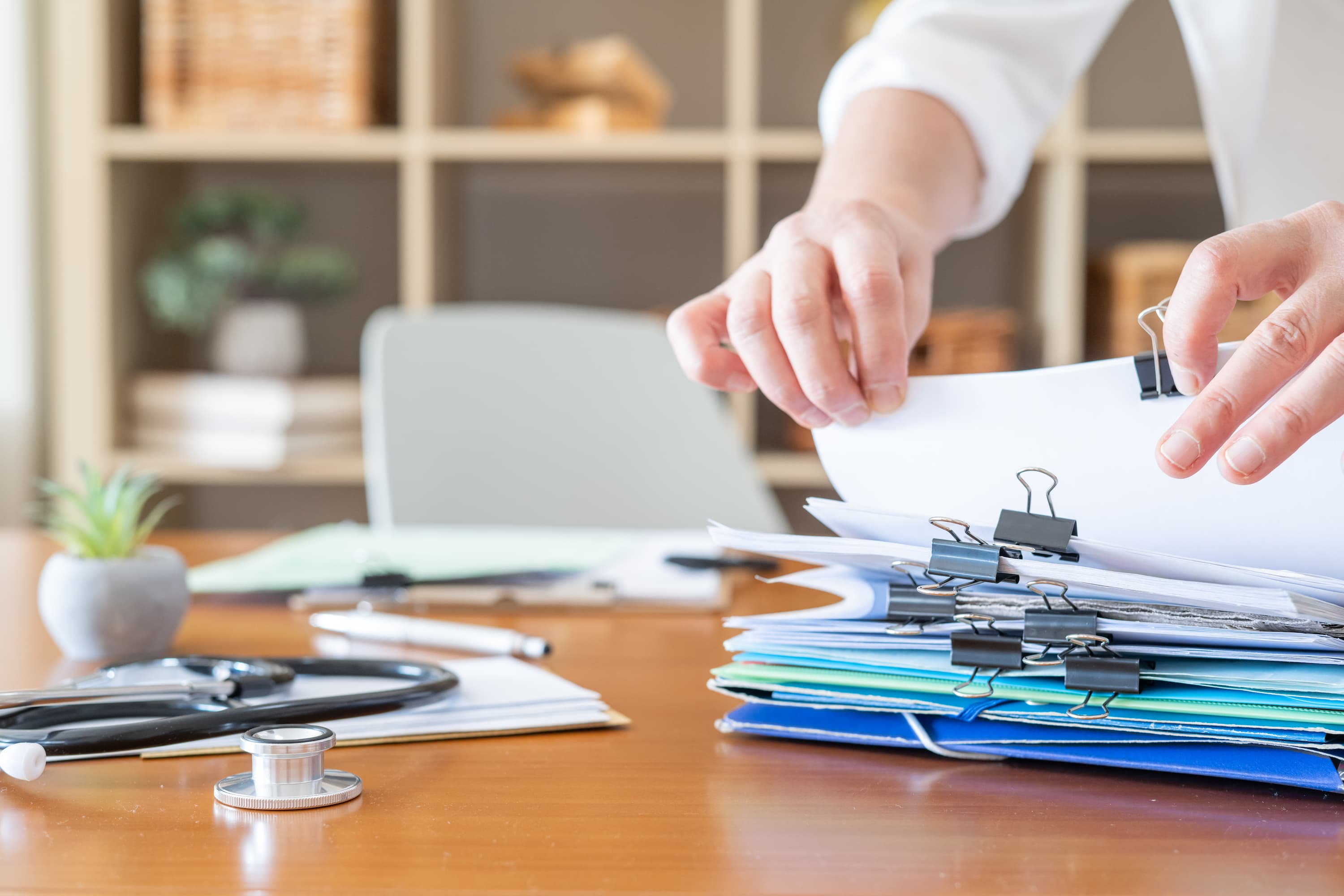 Hands sorting through stacked medical records and paperwork on a desk beside a stethoscope
